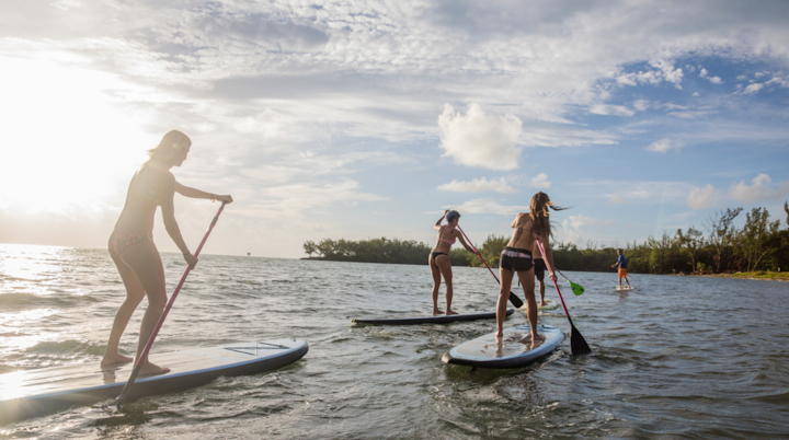 Un groupe de personnes fait du paddle sur une eau calme près d'un rivage tropical, sous un ciel partiellement nuageux au coucher du soleil.