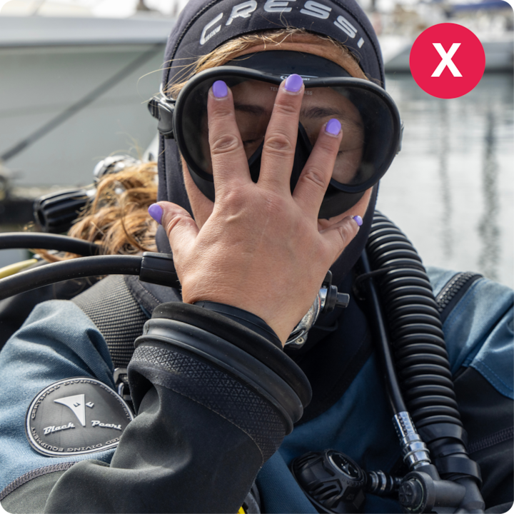 Plan rapproché d'une plongeuse portant son équipement et faisant un geste de la main, avec un port de plaisance et des bateaux flous en arrière-plan.