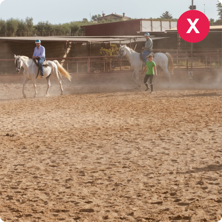 Deux personnes montent des chevaux blancs dans une arène poussiéreuse, guidées par une personne vêtue d'un haut vert qui marche à côté d'elles.