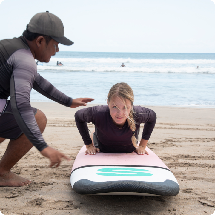 Une personne allongée sur une planche de surf écoute les conseils d'un moniteur. Ils sont sur une plage de sable et l'on aperçoit des vagues en arrière-plan.