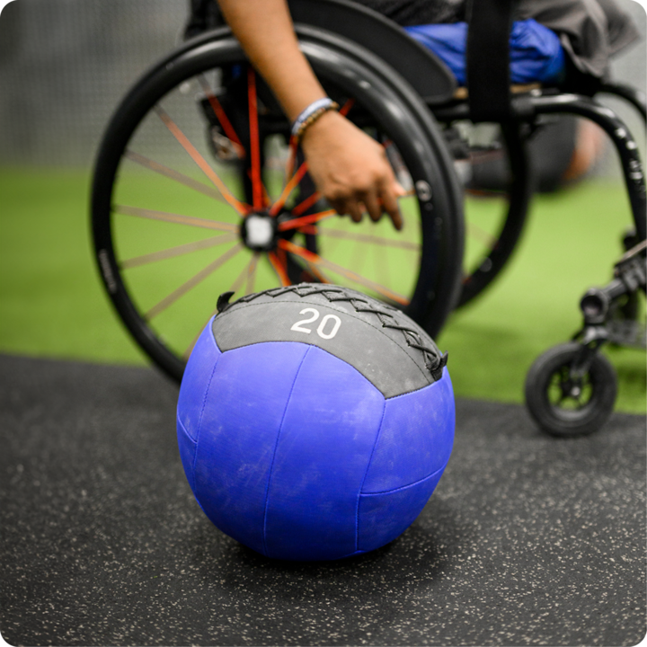 Un médecine ball portant l'inscription « 20 » est posé sur le sol d'une salle de sport. On voit aussi une partie d'un fauteuil roulant et la main d'une personne.