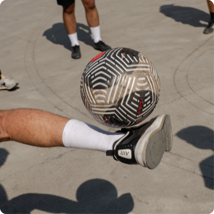 Un ballon de football à motifs semble suspendu au-dessus de la chaussure d'une personne entourée d'autres personnes lors d'une journée ensoleillée.