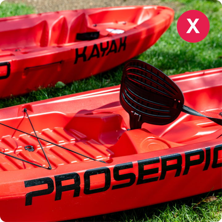 Des kayaks rouges sont posés sur une pelouse. L'un d'eux porte la marque « Proserpic » et une pagaie noire est posée dessus.