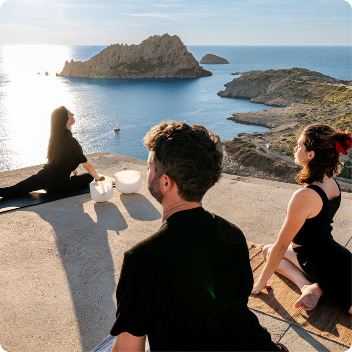 Plusieurs personnes font du yoga sur une terrasse ensoleillée avec une vue panoramique sur l'océan, une île rocheuse et un voilier.