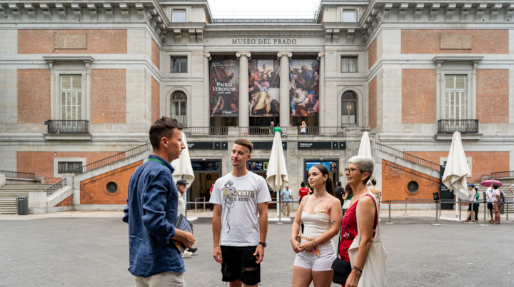 Un groupe de personnes se tient devant le musée du Prado, un musée d'art historique à Madrid, avec de grandes colonnes classiques et des bannières présentant des œuvres d'art.