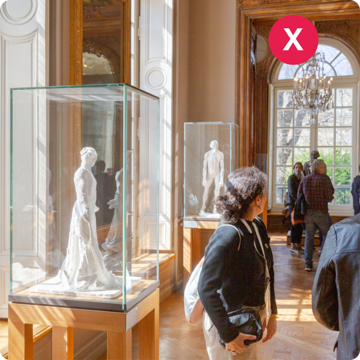Une galerie de musée lumineuse avec des sculptures en marbre dans des vitrines, une femme observant une statue et des visiteurs en arrière-plan.