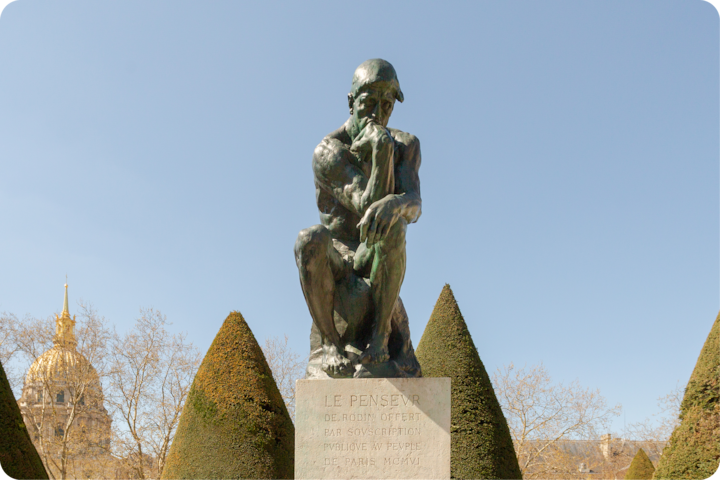 La statue en bronze emblématique « Le Penseur » d'Auguste Rodin, assise sur un piédestal avec une inscription, entourée d'arbres, de haies et d'un dôme doré sous un ciel ensoleillé.