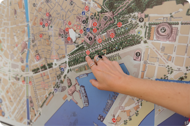 Un gros plan d'une main portant des bagues et pointant un endroit précis sur une carte de ville colorée avec des monuments, des arbres et un bord de mer.