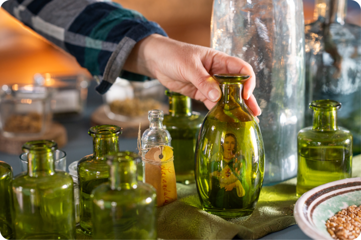 Plan rapproché d'une main sélectionnant une bouteille en verre vert parmi une sélection de verrerie vintage et de bocaux remplis de grains sur une nappe texturée.