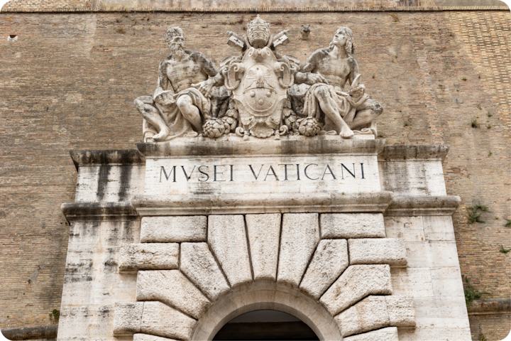 Entrée des musées du Vatican avec des sculptures en pierre ornées de deux personnages assis et un blason papal au-dessus d'une porte arquée.