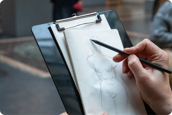Une main dessinant une statue classique d'une femme sur un bloc-notes dans un musée, avec des détails d'arrière-plan flous.
