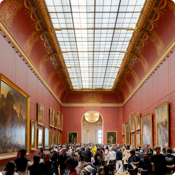 Une salle de musée élégante et bondée, avec des murs rouges somptueusement décorés, des moulures dorées complexes, un toit vitré et de nombreuses grandes peintures historiques.