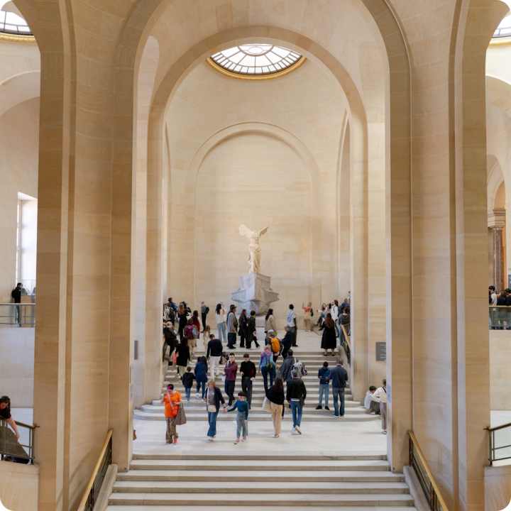 Des visiteurs traversent une grande salle du musée du Louvre et se rassemblent autour de la Victoire de Samothrace.