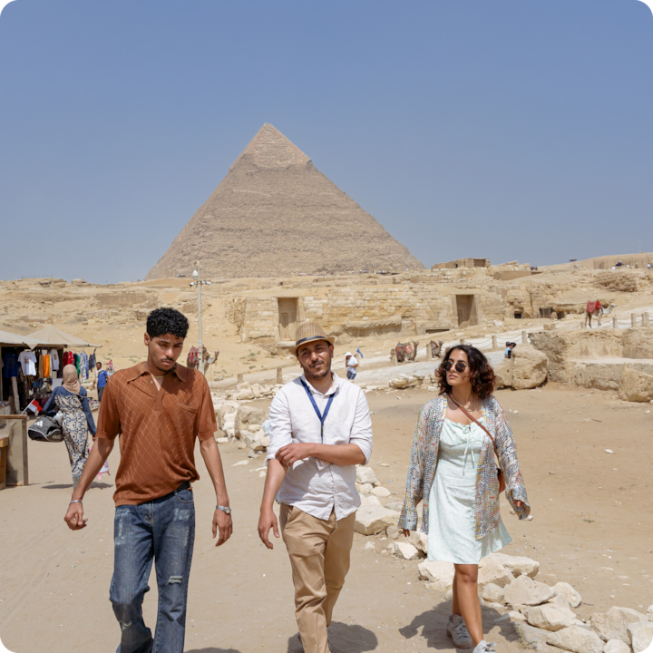 Trois personnes marchent près de la grande pyramide de Gizeh par une journée ensoleillée, entourées de ruines antiques et d'un paysage de sable.
