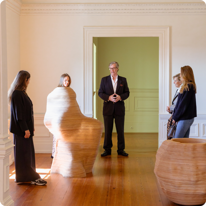 Une salle de musée d'art moderne avec éclairage naturel, où se trouve une sculpture en bois ressemblant à une silhouette assise, entourée de voyageurs qui écoutent un guide.