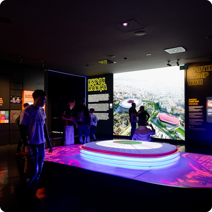 Une salle de musée sombre avec un éclairage coloré, mettant en valeur une table d'exposition futuriste et des visuels de l'Espai Barça, avec des visiteurs lisant des panneaux et observant la projection du stade.
