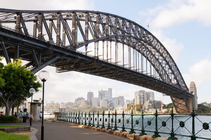 Sydney Harbour Bridge with the city skyline in the background on a sunny day, viewed from a waterfront path in New South Wales.
