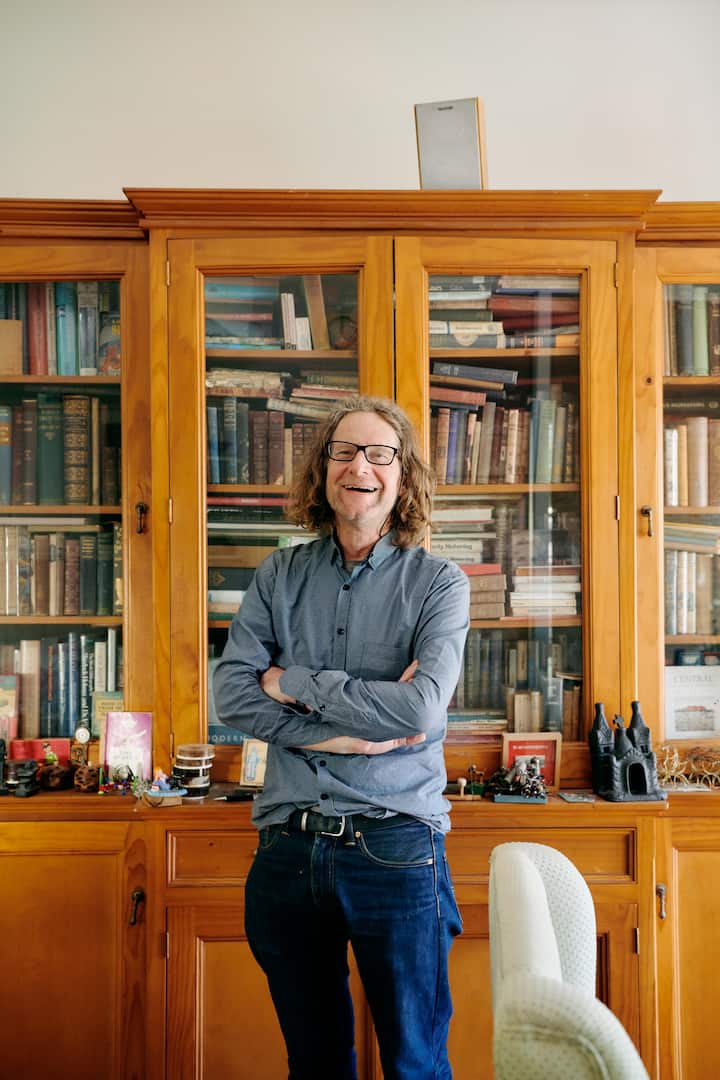 A person standing with arms crossed in front of a wooden bookshelf filled with books and small decorative items, conveying a formal and intellectual setting.