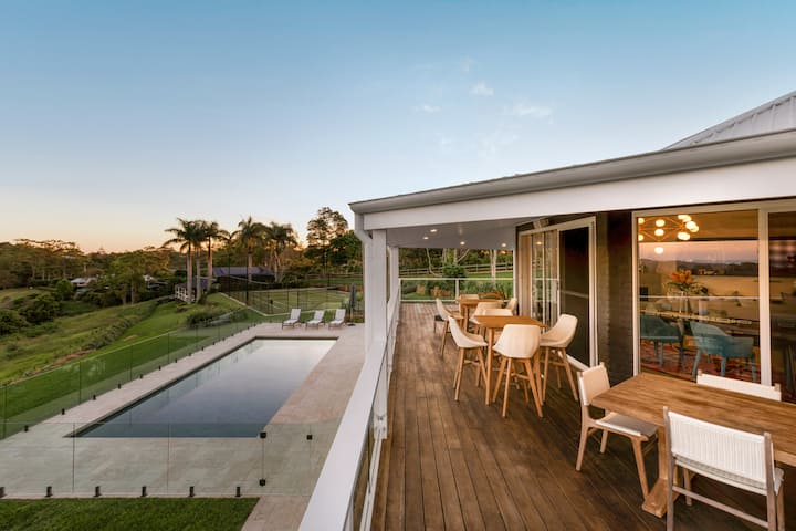 A modern vacation home in New South Wales with an outdoor wooden deck featuring a dining area, a glass-enclosed swimming pool, surrounded by lush greenery and palm trees at sunset.