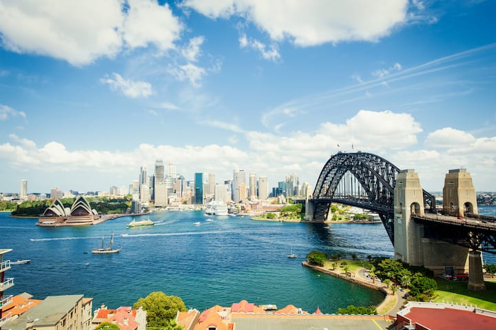 Scenic view of Sydney Harbour featuring the Sydney Opera House on the left, the Harbour Bridge on the right, and the city skyline in the background under a sunny, partly cloudy sky.