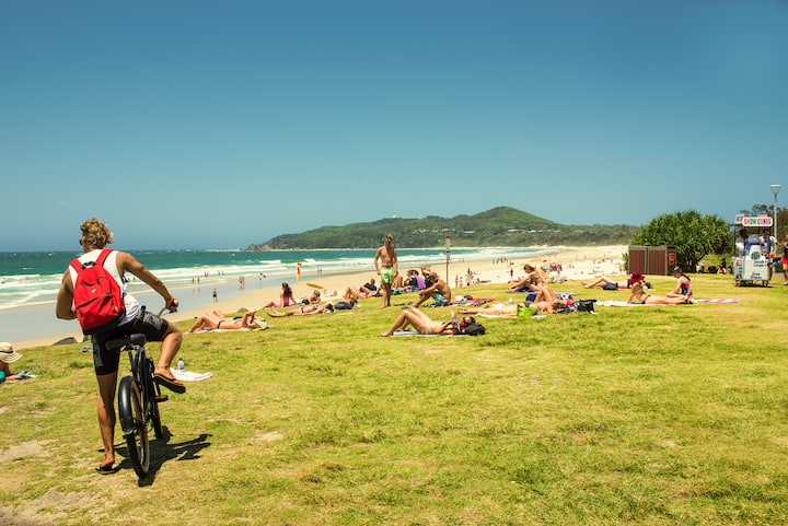 A sunlit beach in New South Wales with people lounging on grassy areas and sandy shores, a cyclist with a red backpack in the foreground.