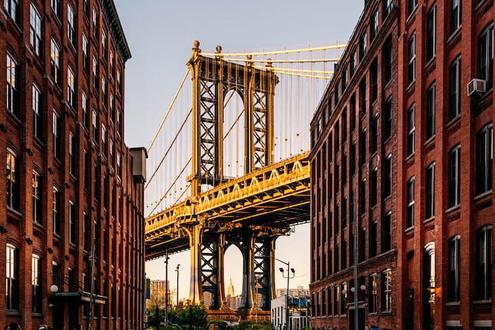 View of the Manhattan Bridge framed by two rows of red brick buildings at sunset, with the Empire State Building visible in the distance.