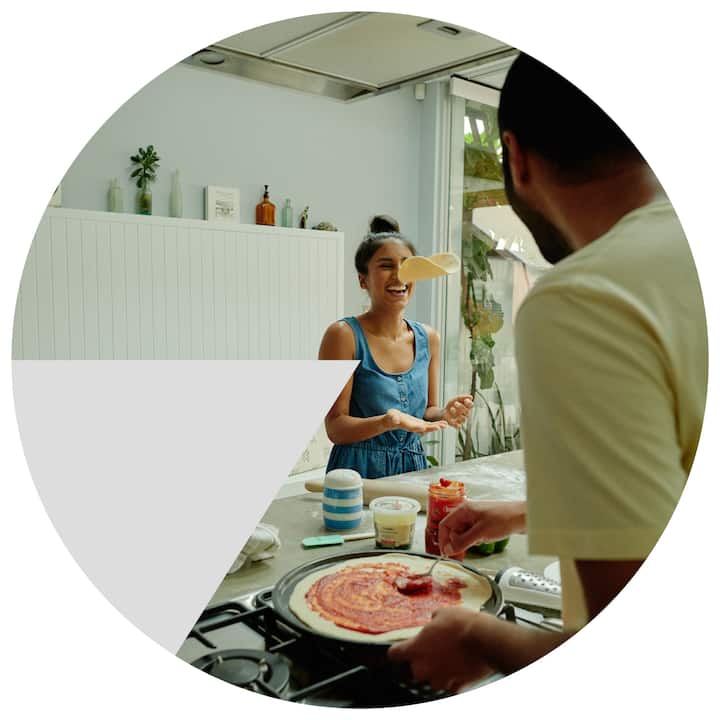 A person spreads tomato sauce on pizza dough at a kitchen counter while another person stands nearby in a light-filled, modern kitchen with greenery visible through the window.