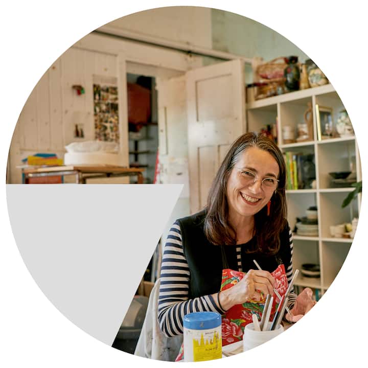 A cozy art studio with shelves filled with pottery and art supplies, featuring a person painting at a table with brushes and colorful jars nearby.