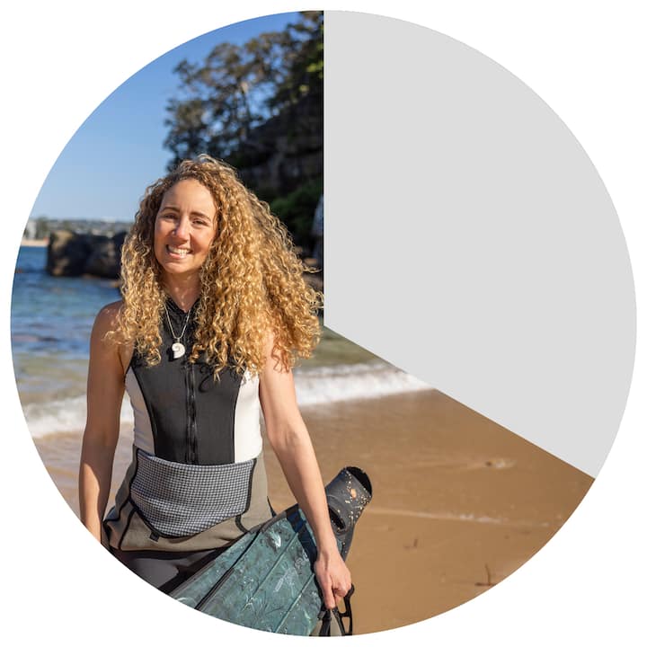 A person holding snorkeling gear stands on a sandy beach with calm water and rock formations in the background under a clear blue sky, representing outdoor recreation in New South Wales.