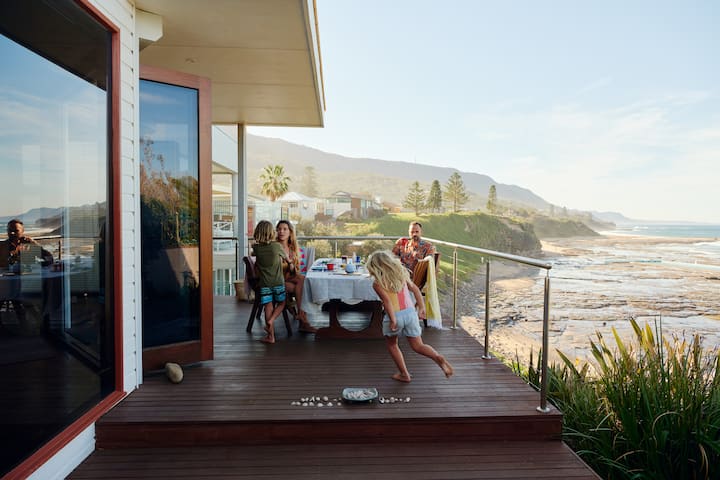 A family seated at a table on a wooden deck overlooking a scenic coastline in New South Wales, with a child running towards the table. The ocean, coastal cliffs, and houses are visible in the background.