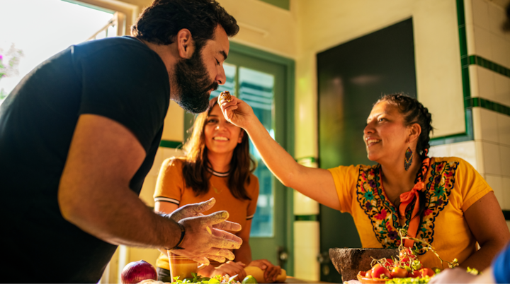 Les participants se rassemblent autour d'une table avec des produits frais tandis qu'un hôte vêtu d'une tenue colorée offre des conseils culinaires dans une cuisine lumineuse et accueillante.