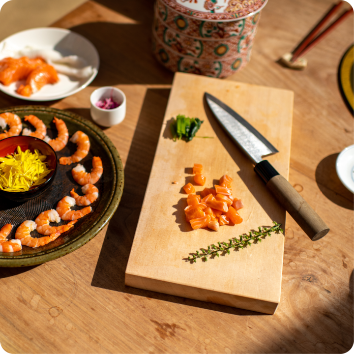 Une planche à découper en bois avec du saumon en dés, un brin de thym et un couteau, à côté d'une assiette de crevettes, de fleurs comestibles et de petits bols de garnitures.