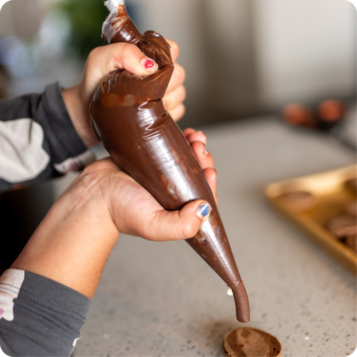 Une personne presse une poche à douille pour déposer du chocolat sur une coque de macaron, avec des ustensiles de pâtisserie visibles sur le comptoir.