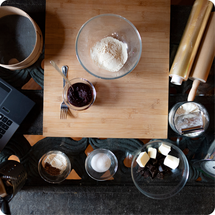 Vue plongeante d'une planche à découper en bois avec de la farine, de la confiture et divers ingrédients de pâtisserie comme du beurre, du sucre et du chocolat, entourée d'ustensiles de cuisine, notamment des rouleaux à pâtisserie et un ordinateur portable.