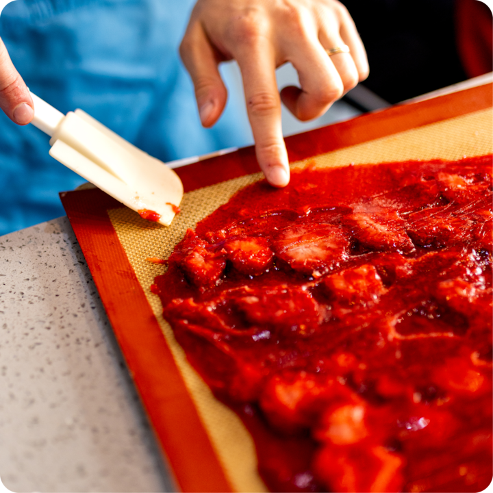 Des mains étalent un mélange de fraises rouge vif sur un tapis de cuisson à l'aide d'une spatule, dans le cadre d'une préparation culinaire.