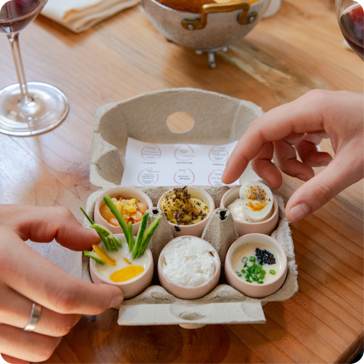 Des mains rangent six plats miniatures uniques dans une boîte à œufs. Les ingrédients de l'expérience culinaire, des herbes, du caviar et des sauces, sont colorés.