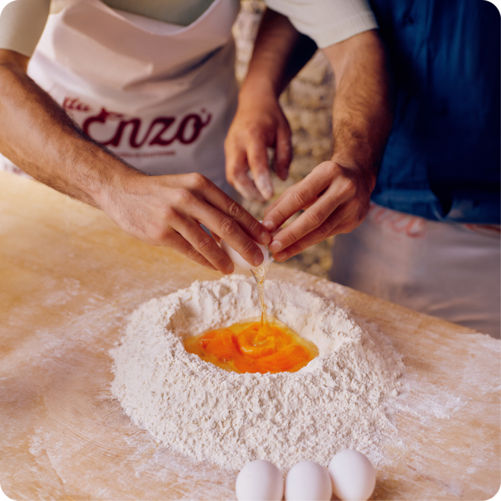 Deux personnes cassent un œuf dans un tas de farine sur une surface en bois, préparant une pâte ensemble.