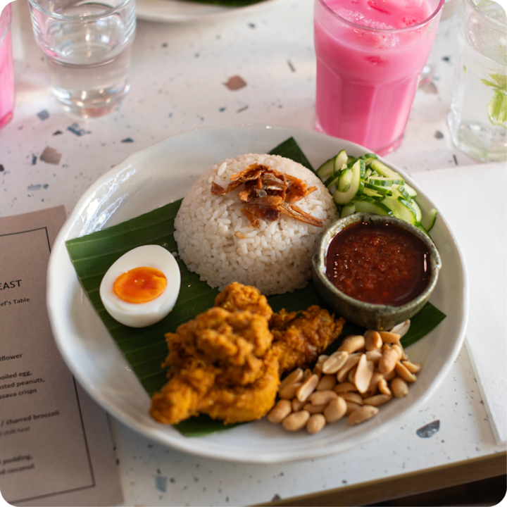 Un gros plan d'un plat coloré composé de riz blanc, de sambal, de poulet frit, de cacahuètes, de tranches de concombre et d'un œuf mollet, placé sur une feuille de bananier. Des boissons roses et transparentes sont visibles en arrière-plan.