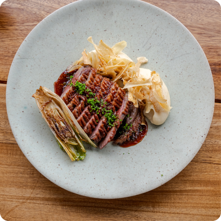 Un plat élégamment présenté, composé d'un steak saisi avec un filet de sauce, de pommes de terre frites croustillantes, d'endive rôtie et de purée de pommes de terre sur une assiette aux tons neutres.