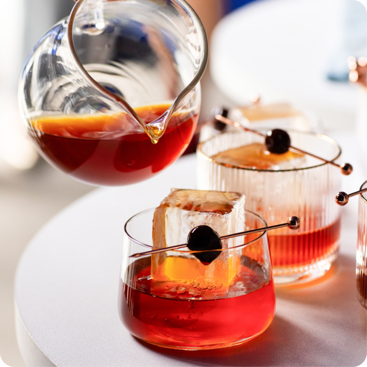 Gros plan d'une carafe en verre versant un liquide rouge vif dans un verre à cocktail avec un gros glaçon et une cerise sur pique.