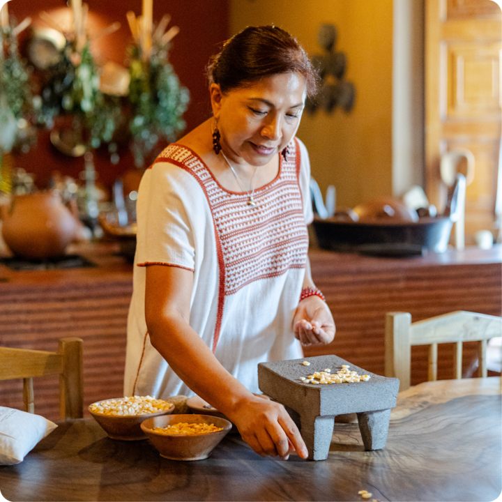 Plan rapproché de mains travaillant avec un broyeur en pierre pour écraser des grains de maïs, entourées de bols de maïs dans une cuisine chaleureuse et bien éclairée aux tons naturels, décorée de plantes suspendues.