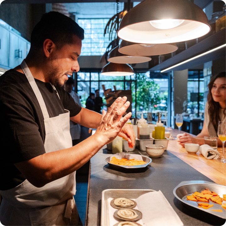 Un chef expérimenté apporte la touche finale à un plat dans une cuisine chaleureusement éclairée, mettant en valeur son art culinaire avec des ingrédients frais, tandis que les convives sont assis au comptoir dans une atmosphère décontractée et accueillante.