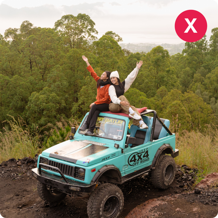 Deux personnes assises sur un 4x4 turquoise, les bras levés, entourées d'une forêt verdoyante et surplombant une ville au loin.