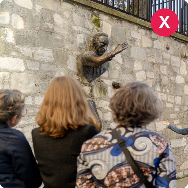 Des visiteurs admirent une sculpture en bronze encastrée dans un mur de pierre représentant un homme qui tend la main, guidés par un hôte qui présente l'œuvre.
