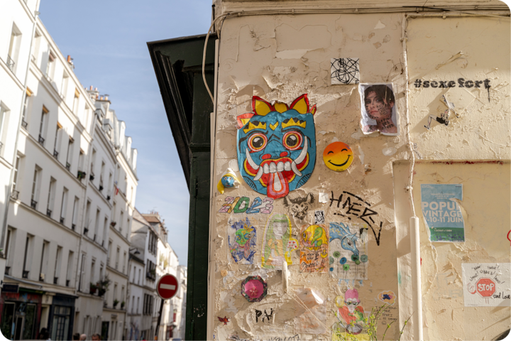 Un gros plan d'un mur orné de street art, avec un dessin ressemblant à un masque aux yeux rouges et à la langue tirée, entouré de graffitis et d'affiches, dans un paysage urbain lumineux.