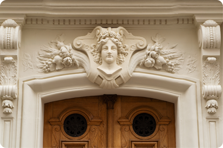 Une arche en pierre blanche sculptée au-dessus d'une double porte en bois ornée de détails floraux complexes et d'un écusson sculpté au centre.
