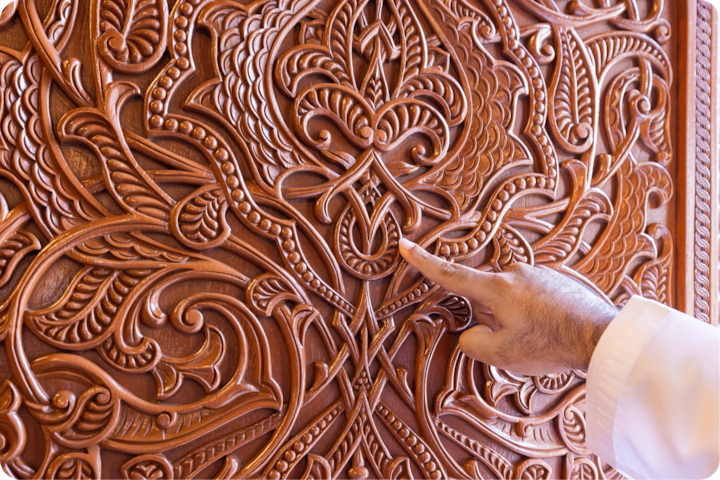 Plan rapproché d'une main pointant vers des sculptures en bois élaborées avec des motifs floraux et ornementaux tourbillonnants, mettant en valeur l'art traditionnel.