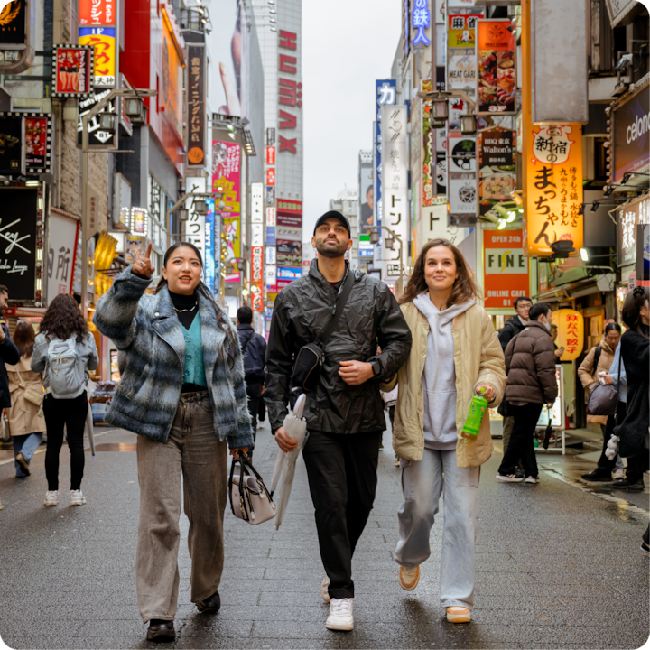 Une rue animée avec des publicités lumineuses et des piétons, avec trois personnes marchant au premier plan, explorant le quartier.