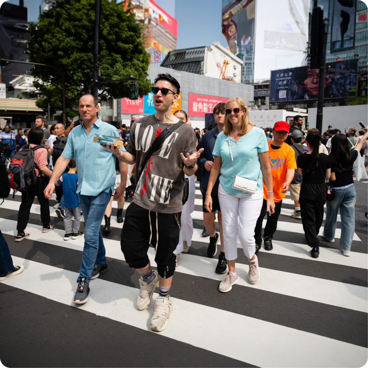 Une scène urbaine animée montrant des personnes traversant un passage piéton, de grands panneaux d'affichage et une architecture moderne en arrière-plan.