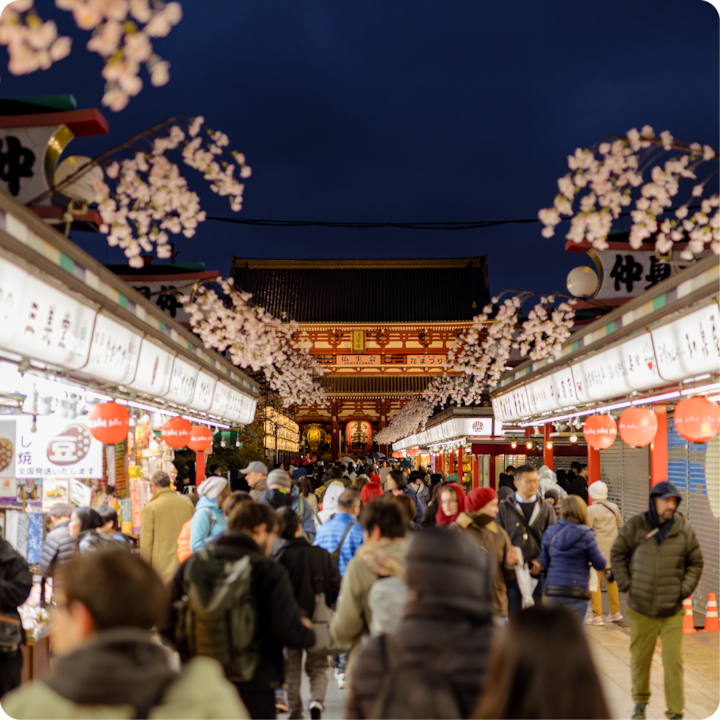 Un marché en plein air animé devant un temple japonais traditionnel la nuit, entouré de cerisiers en fleurs et d'étals illuminés.
